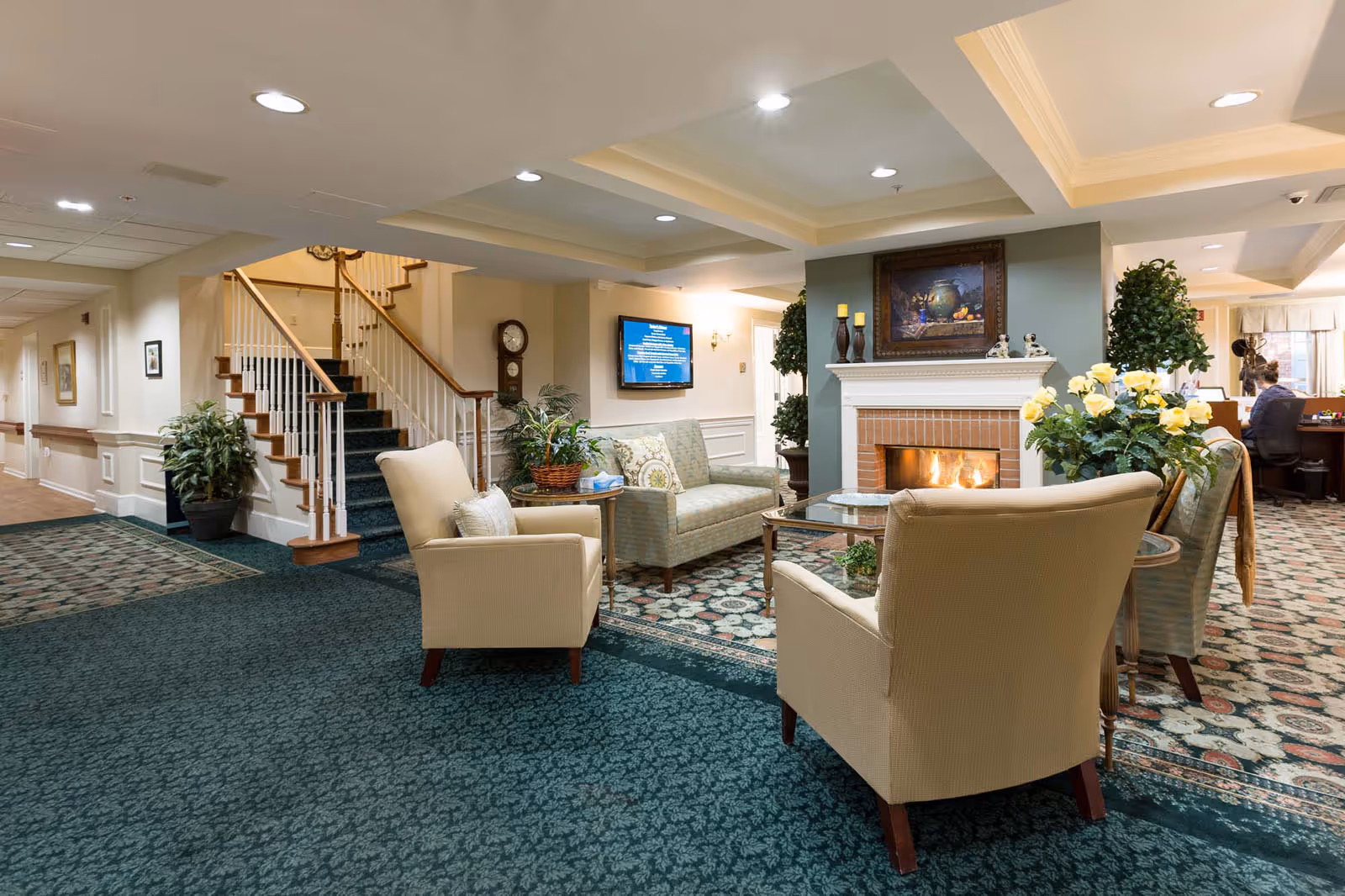 A cozy senior living facility common area with a fireplace, two beige armchairs, a patterned sofa, a glass coffee table, and decorative plants. A staircase with wooden handrails is visible in the background, along with a wall-mounted TV and a grandfather clock. The room is warmly lit with recessed ceiling lights.