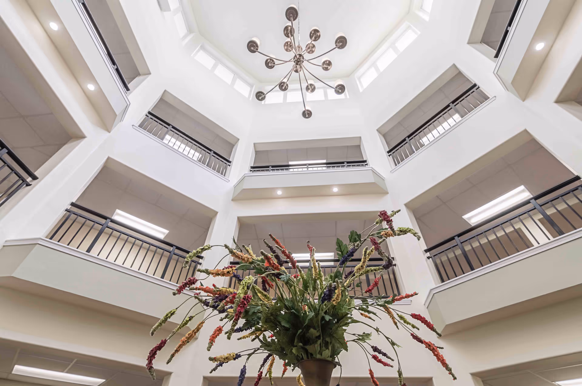 View looking up inside a multi-story atrium with white walls, black railings on balconies, and a modern chandelier hanging from the ceiling. A large floral arrangement with colorful flowers is visible at the bottom center of the image.