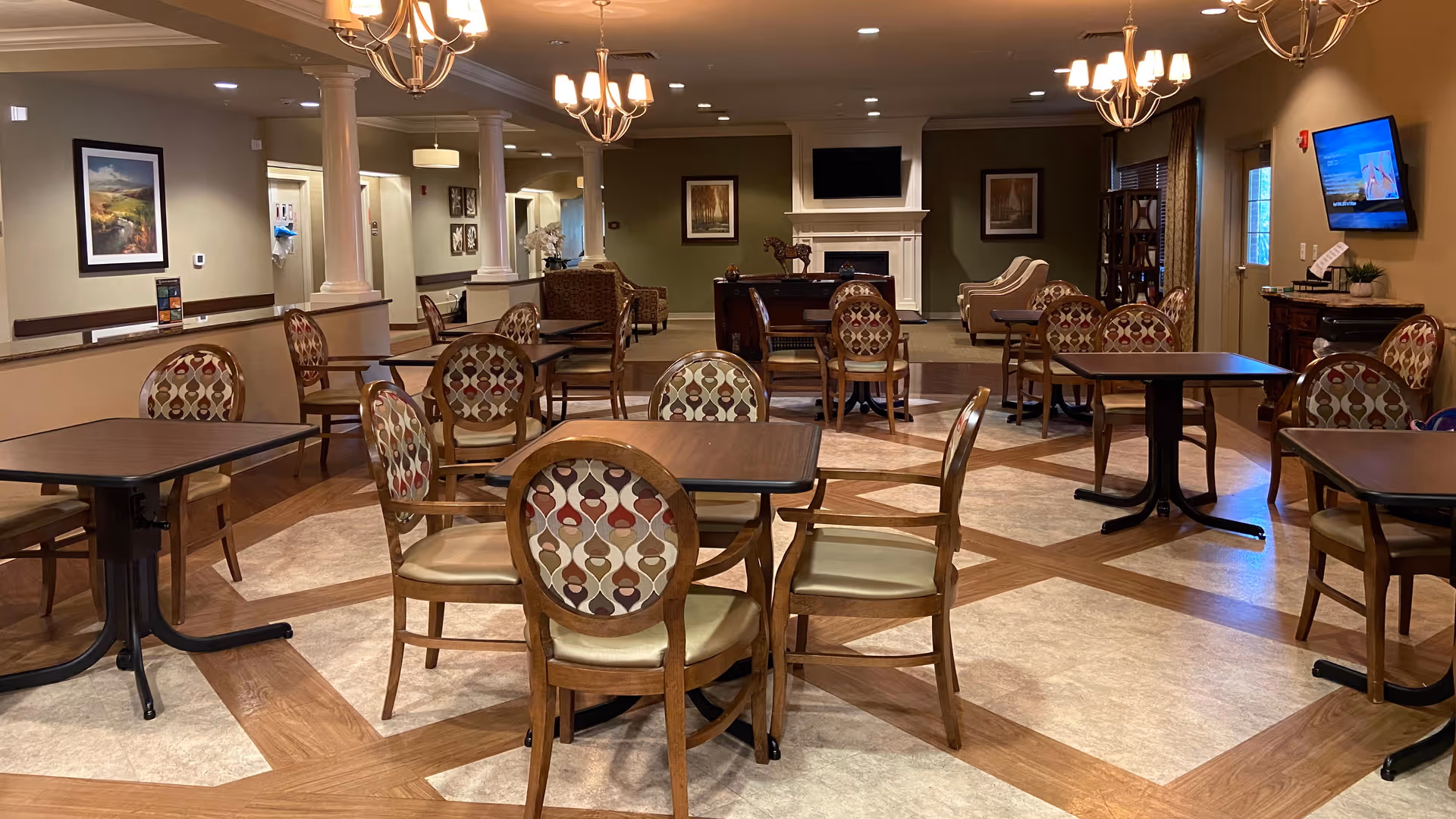 A spacious, well-lit common area with multiple wooden tables and patterned cushioned chairs arranged neatly. The room features a decorative fireplace with a TV mounted above it, framed artwork on the walls, and chandeliers hanging from the ceiling. There is a mix of carpet and wood flooring, and a window with curtains on the right side.