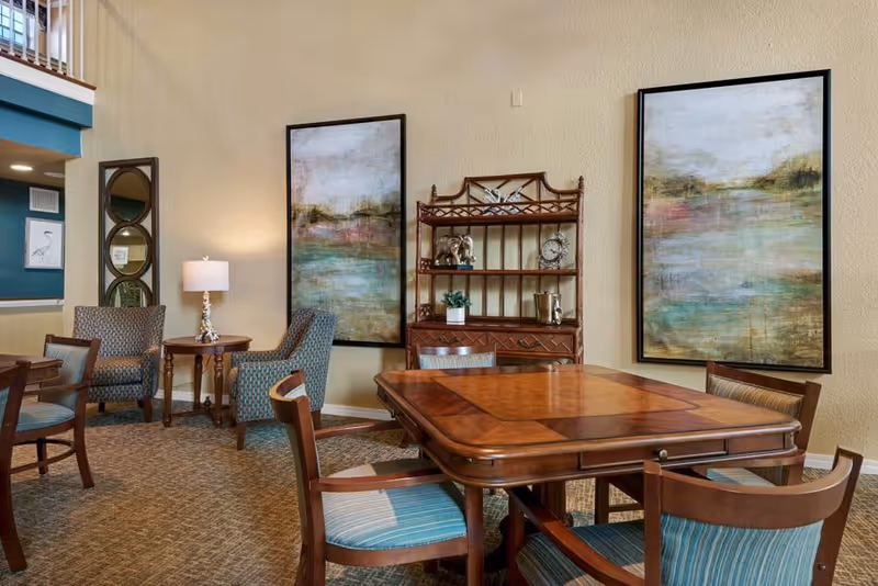 A cozy interior room with a wooden table surrounded by six chairs with blue striped cushions. Two large abstract landscape paintings hang on the beige walls. There is a wooden shelving unit with decorative items including a clock and a small plant. To the left, there are two upholstered armchairs with a small round table and a lamp between them. The room has carpeted flooring and a high ceiling with a partial view of a second floor railing.