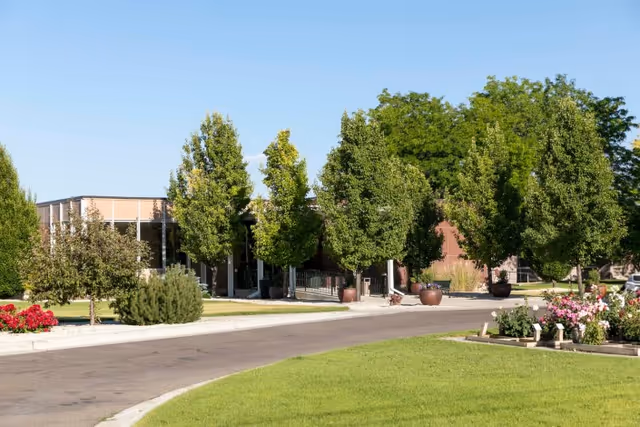 Landscaped driveway and entrance of a rehabilitation and care facility with trees, potted planters, and a manicured lawn.
