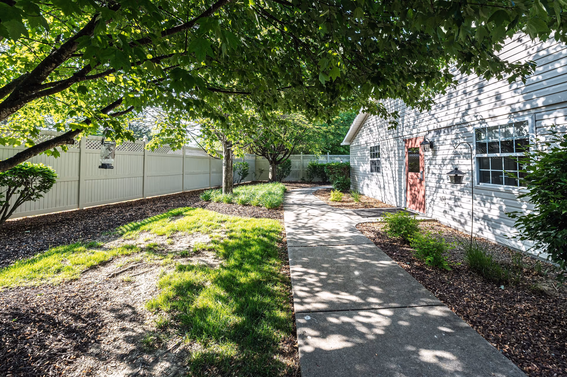 A shaded outdoor walkway next to a white building with a red door, surrounded by green trees, bushes, and a white fence under bright sunlight.