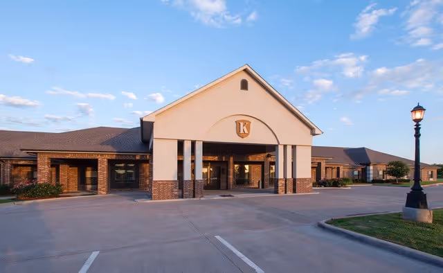Exterior front view of Kemp Care Center building with a large covered entrance, brick and beige facade, parking spaces in front, and a street lamp on the right side under a partly cloudy sky.
