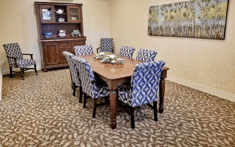 A dining room with a wooden table surrounded by eight patterned upholstered chairs, a china cabinet, and framed wall art.