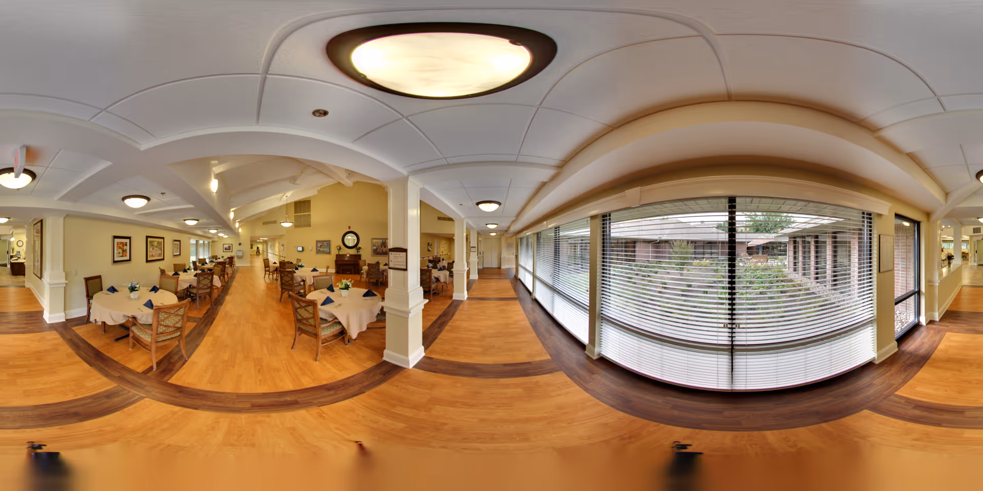 A spacious dining room with multiple round tables covered with beige tablecloths and set with blue napkins. Wooden chairs surround the tables. The room has light yellow walls adorned with framed artwork and a large window with blinds overlooking an outdoor garden area. The floor is wooden with a darker wood inlay pattern. Ceiling lights provide warm illumination.