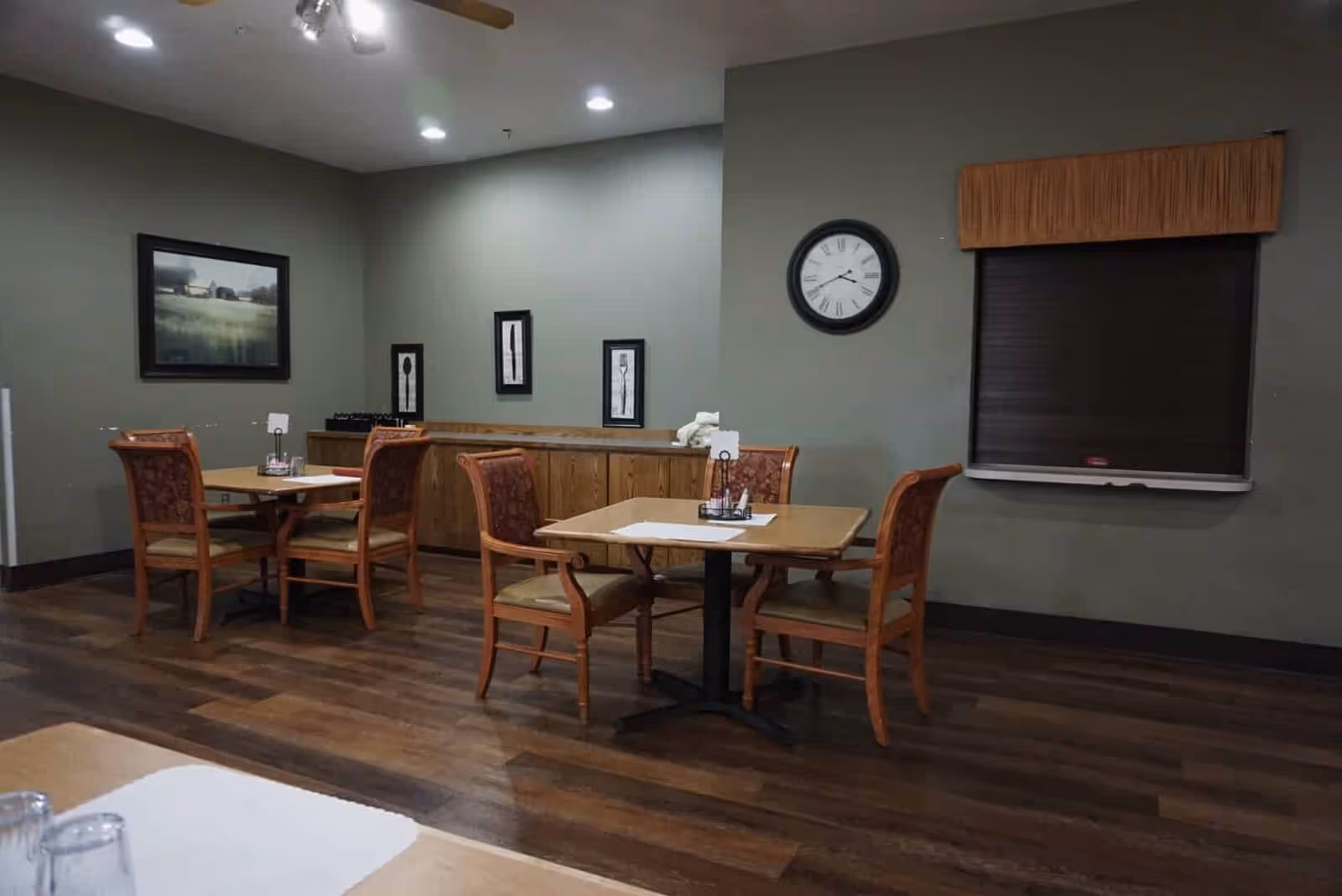 Interior view of a senior living facility dining room with wooden tables and chairs arranged on a wooden floor. The walls are painted green and decorated with framed artwork and a clock. A window with a closed blind is visible on the right side.