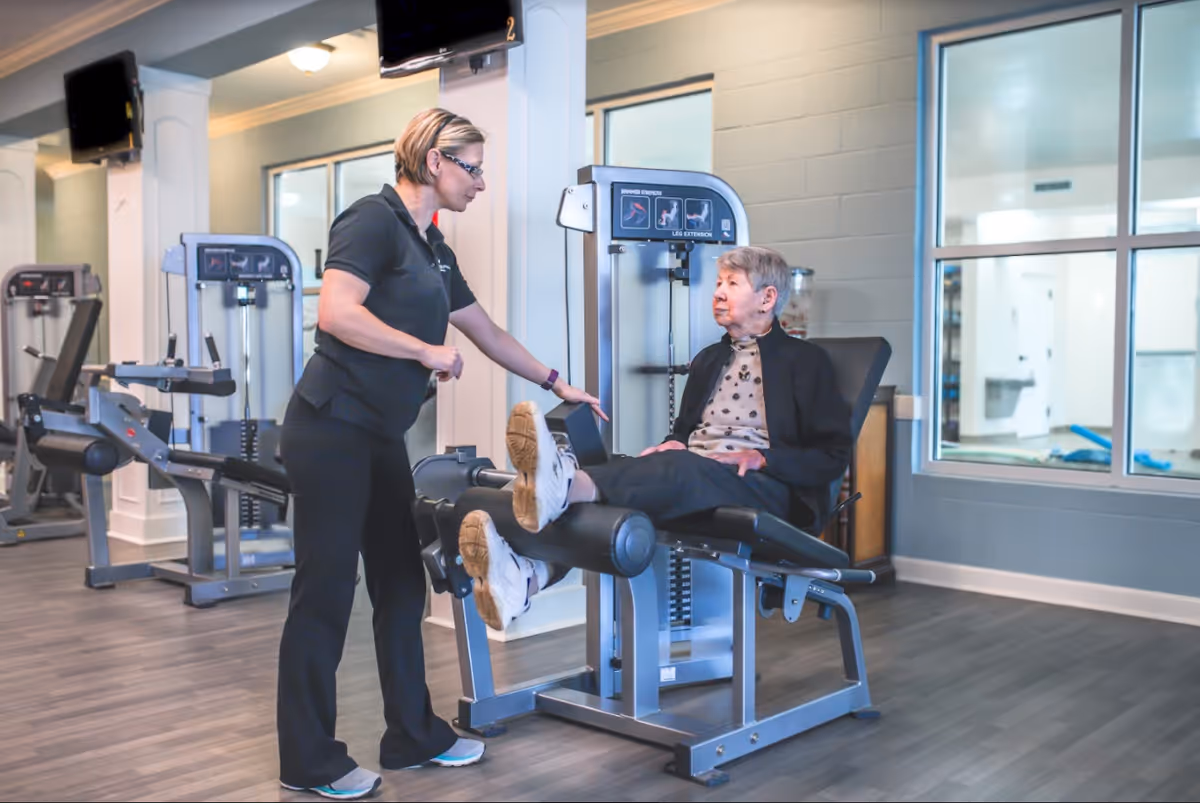 A senior woman is seated on a leg extension exercise machine in a fitness room, while a female instructor or physical therapist stands beside her, providing guidance. The room has exercise equipment and large windows.
