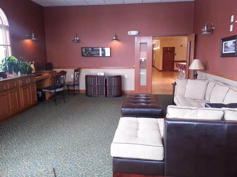 A cozy living room area with a beige sectional sofa and a dark brown ottoman in the center. The room has a green patterned carpet, reddish-brown walls with wooden trim, and several wall-mounted lights. There is a window with blinds and plants on a wooden cabinet to the left, a small desk with chairs, and a doorway leading to a hallway with bar stools visible in the distance.
