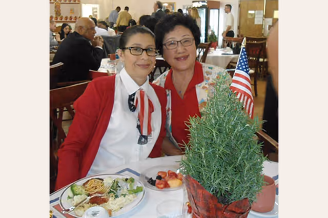 Two women sitting at a dining table in a restaurant or dining room, smiling at the camera. The table has plates with food, including salad and fruit, and a small American flag is placed in a potted plant centerpiece.