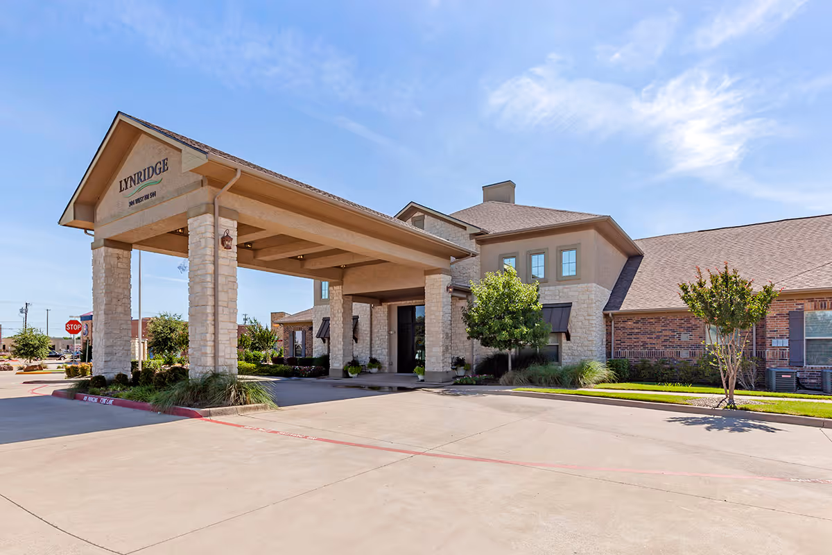 Exterior view of Lynridge of Murphy Assisted Living & Memory Care building with a covered entrance supported by stone pillars, surrounded by landscaping and a clear blue sky.