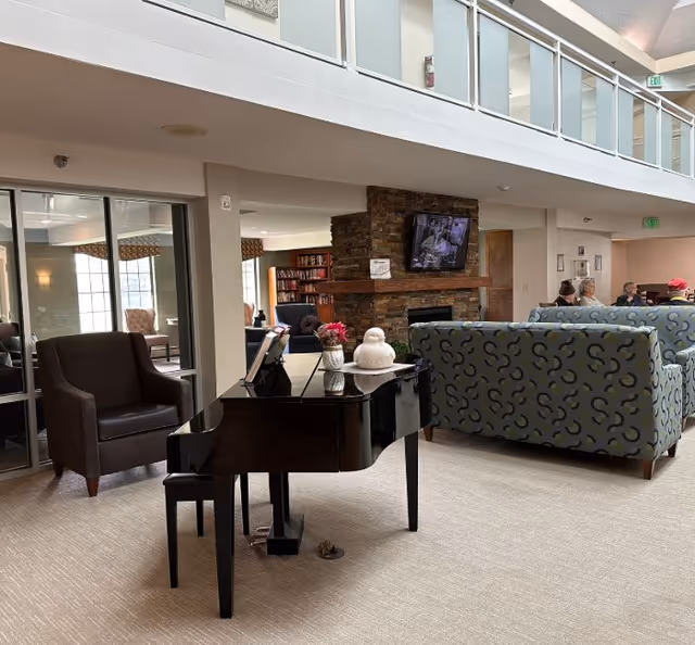 A senior living facility common area featuring a black grand piano with a small bench, a brown armchair, patterned blue sofas, and a stone fireplace with a mounted TV above it. There are people sitting on the sofas in the background, and bookshelves are visible near the fireplace. The area has a high ceiling with a second-floor balcony and glass railings.