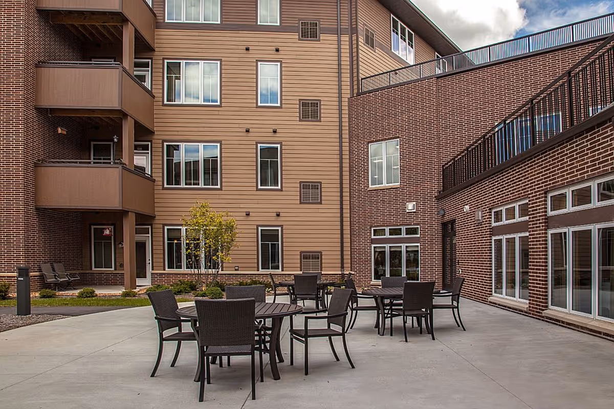 Outdoor courtyard with round tables and chairs in front of a multi-story brick and siding senior living building.