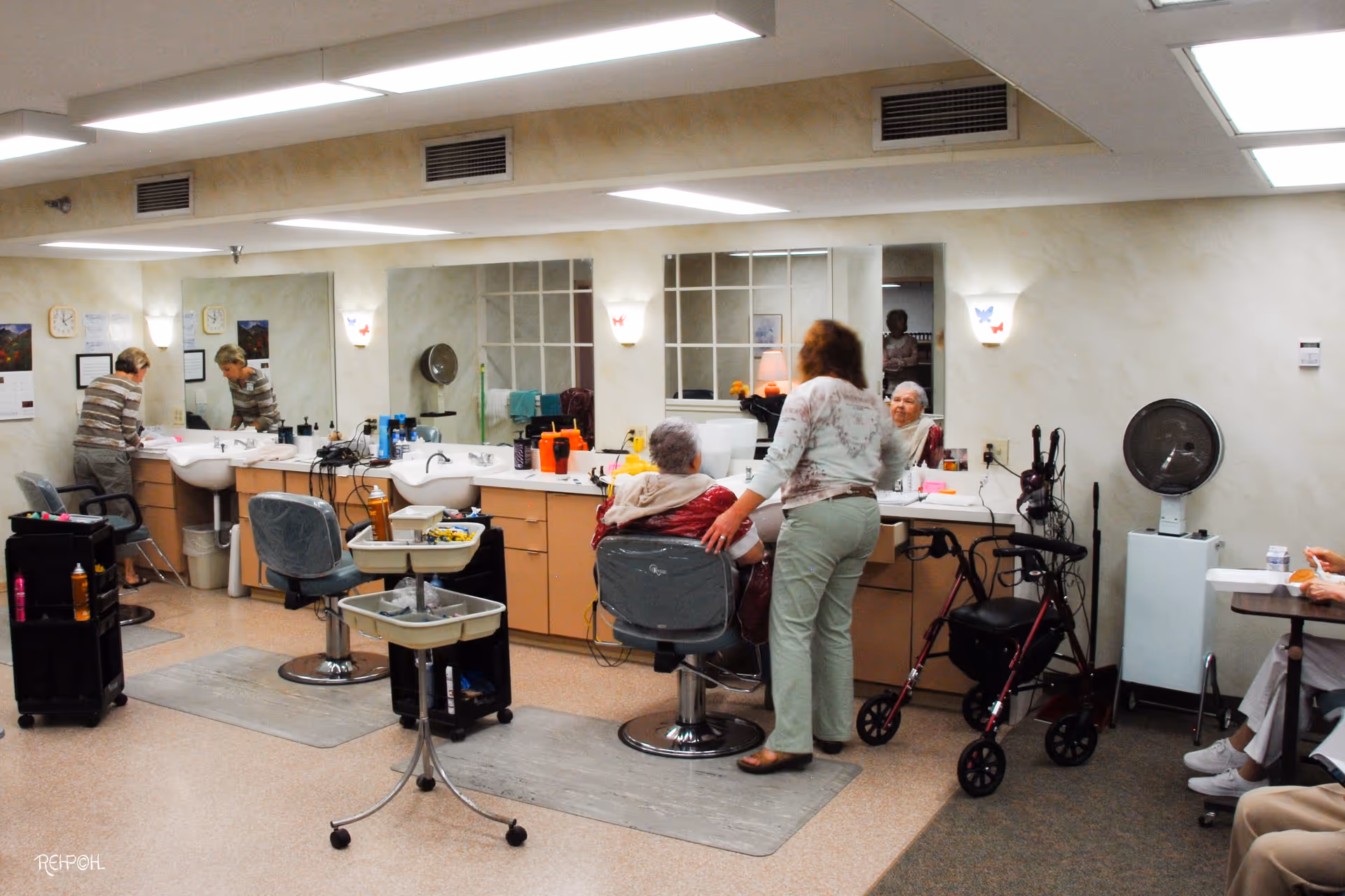 Interior of a senior living facility salon area with two women, one sitting in a salon chair and the other standing beside her. There are mirrors, sinks, salon chairs, and hair styling equipment visible. A walker is placed near the seated woman, and another person is partially visible sitting at a table on the right side.
