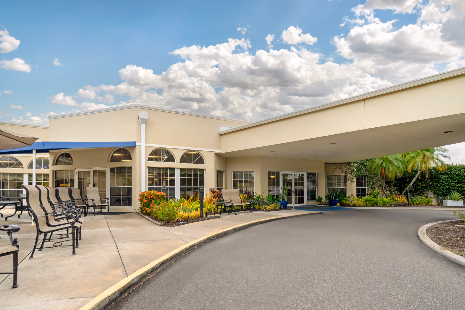 Covered entrance and drop-off area of a senior living facility with outdoor seating, landscaping, and a curved driveway under a partly cloudy sky.