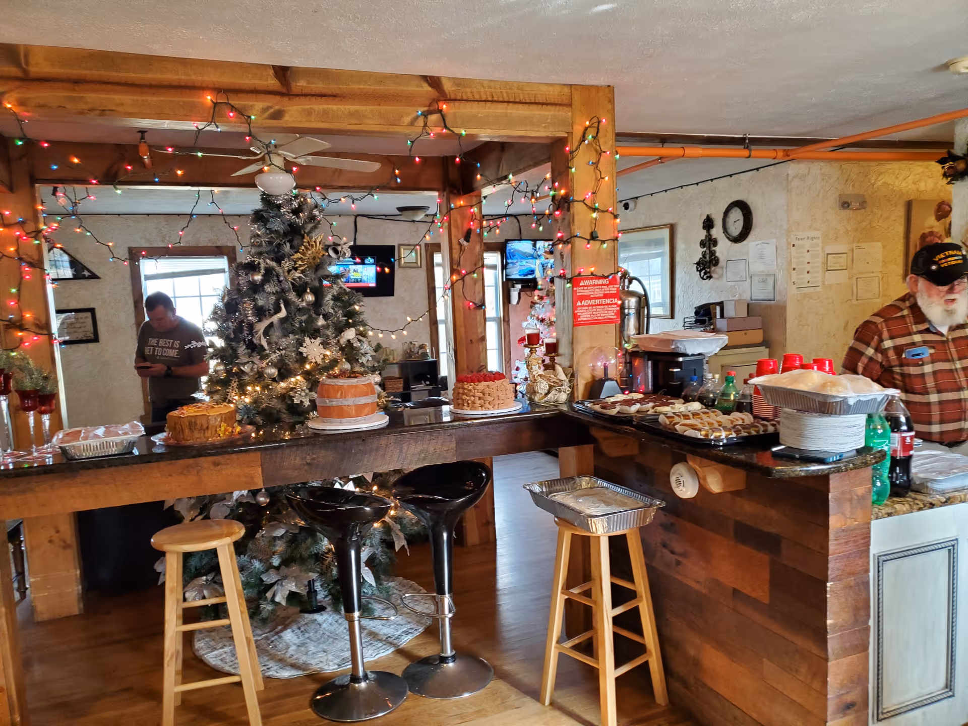 Interior of a cozy room decorated with Christmas lights and a decorated Christmas tree. A wooden counter is set up with various cakes, cookies, and drinks. There are stools around the counter, and two men are present in the background, one wearing a plaid shirt and a hat, and the other looking at a phone.