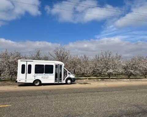 A white Nazareth House of Fresno shuttle bus parked on the roadside in front of a blooming orchard under a blue sky.