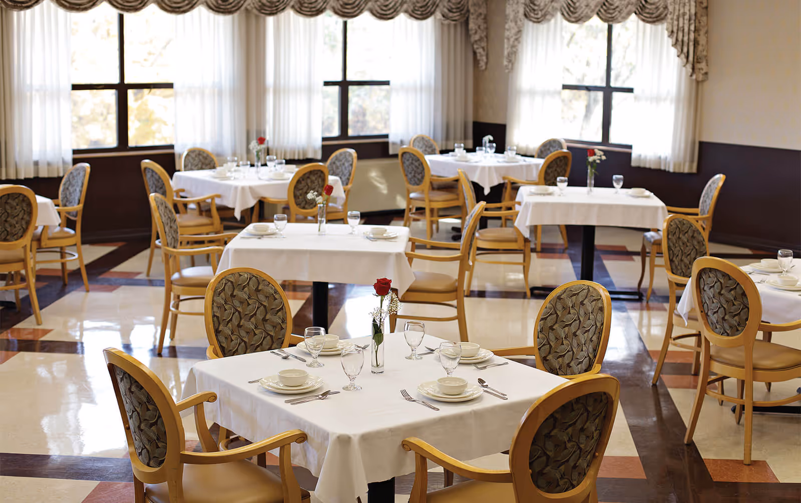 A bright dining room with multiple tables covered in white tablecloths, each set with plates, bowls, glasses, and silverware. Each table has a small vase with a single red rose and some white flowers. The room has large windows with sheer curtains and patterned valances, and wooden chairs with cushioned seats and backs featuring a leaf pattern.