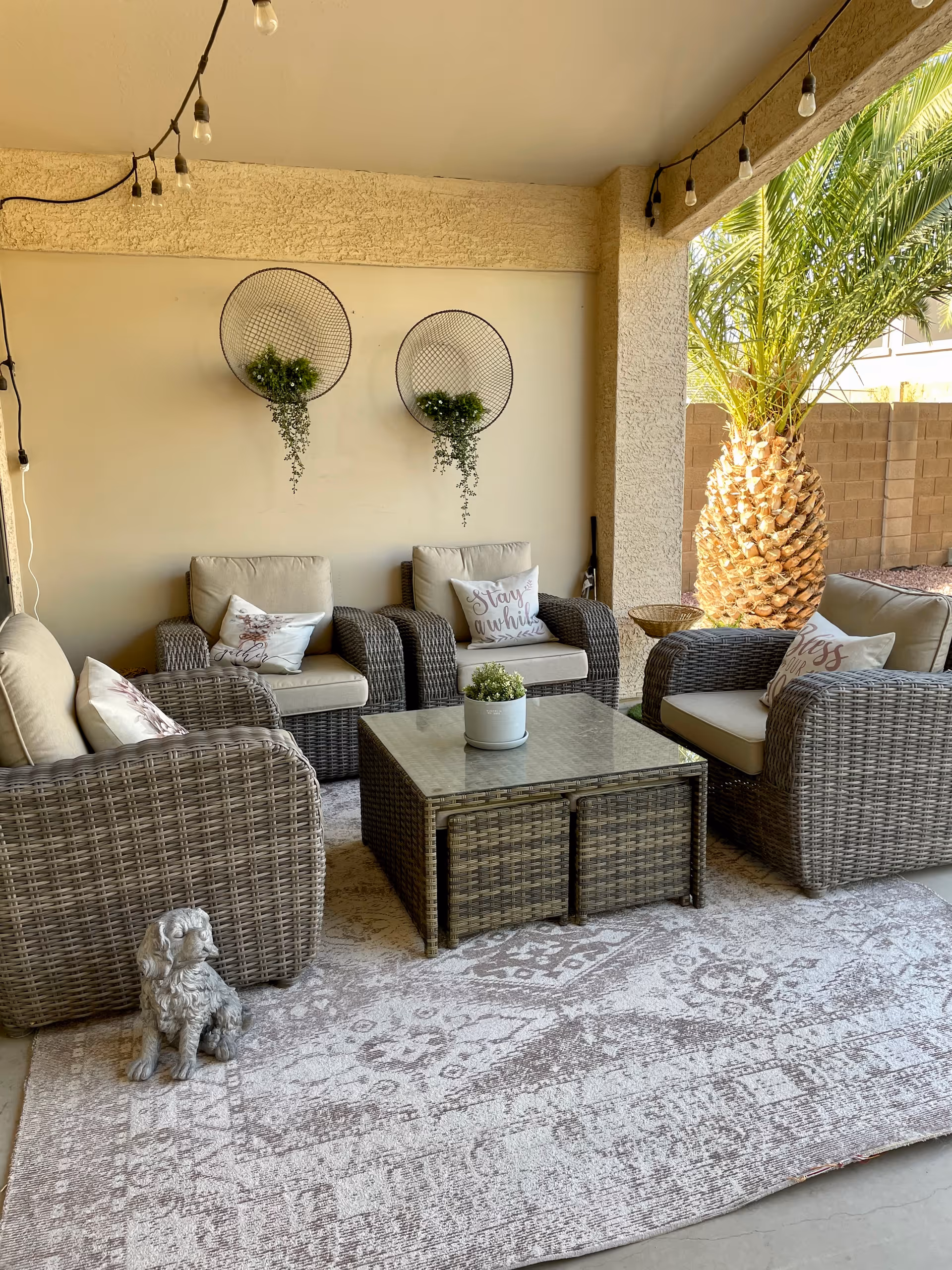 Covered patio seating area with wicker armchairs and cushions arranged around a central wicker coffee table on a patterned rug, string lights overhead and a palm tree visible outside.