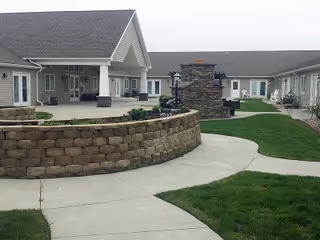 Outdoor courtyard area of a senior living facility with a stone wall, paved walkways, green grass, and a stone fireplace. The building surrounds the courtyard with multiple windows and doors visible.