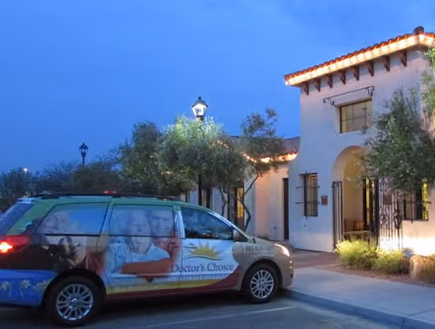 Evening view of the exterior of Doctor's Choice Assisted Living facility with a decorated van parked in front. The building has white walls, a tiled roof, and outdoor lighting. Trees and shrubs are visible near the entrance.