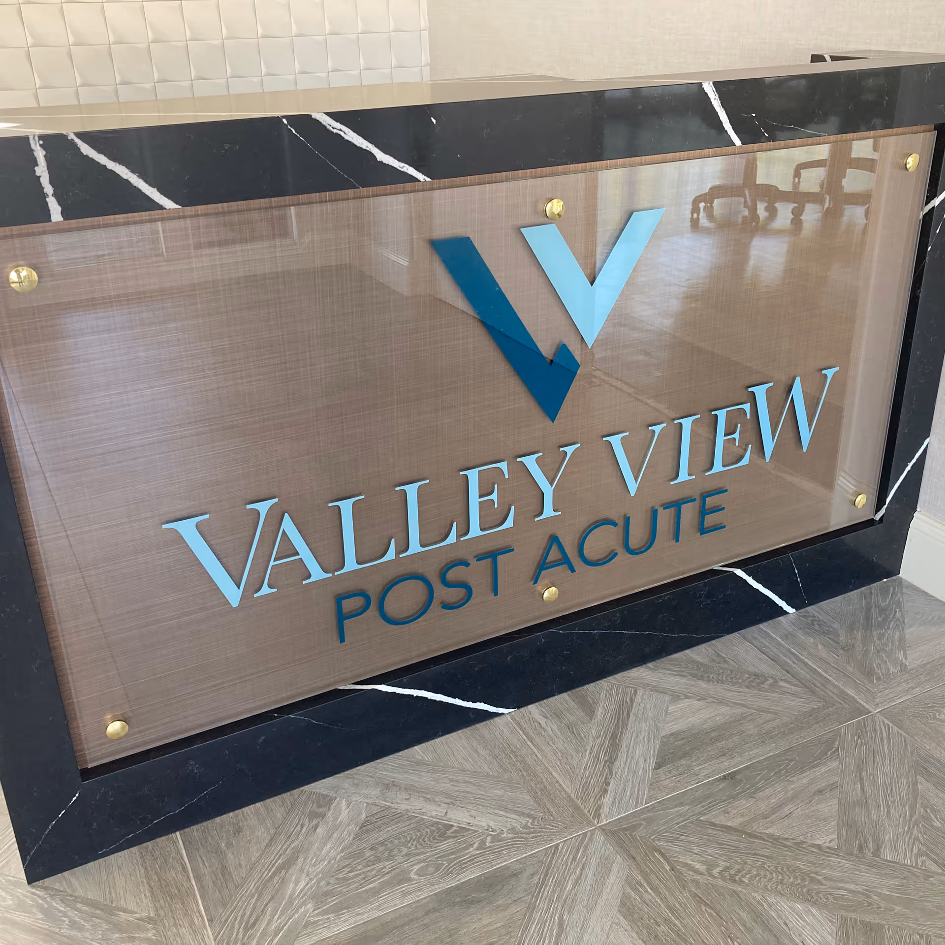 A close-up view of a sign for Valley View Post Acute, mounted on a black marble surface with white veining, with a wooden textured background and gold screws at the corners.