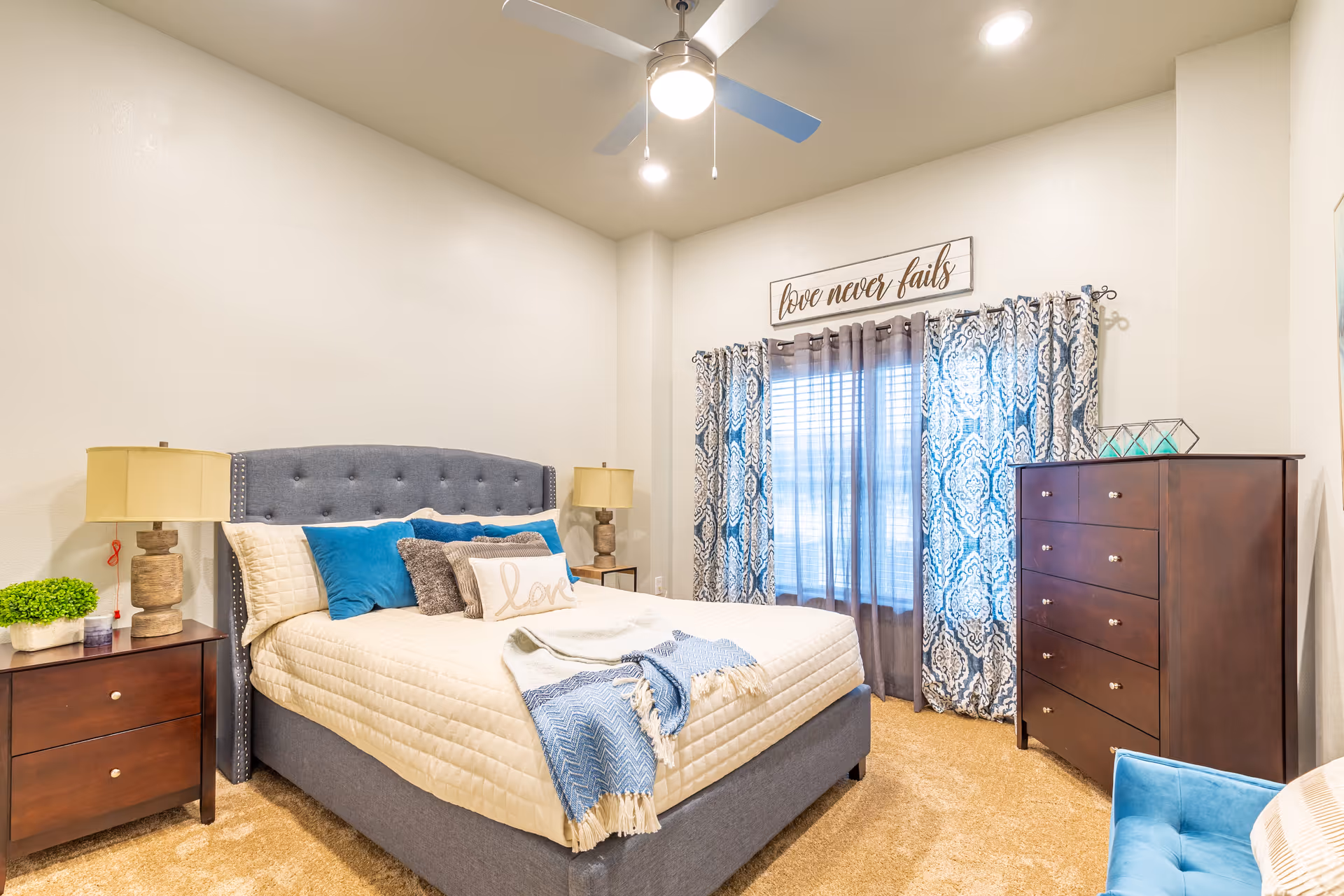 A cozy bedroom with a gray upholstered bed featuring cream-colored bedding and blue accent pillows. There are two wooden nightstands with lamps on either side of the bed. A window with patterned blue and white curtains and sheer gray drapes is on the far wall, above which hangs a sign that reads 'love never fails'. A dark wooden dresser is positioned to the right of the window, and a blue upholstered chair is partially visible in the corner. The room has beige carpeting and a ceiling fan with a light.