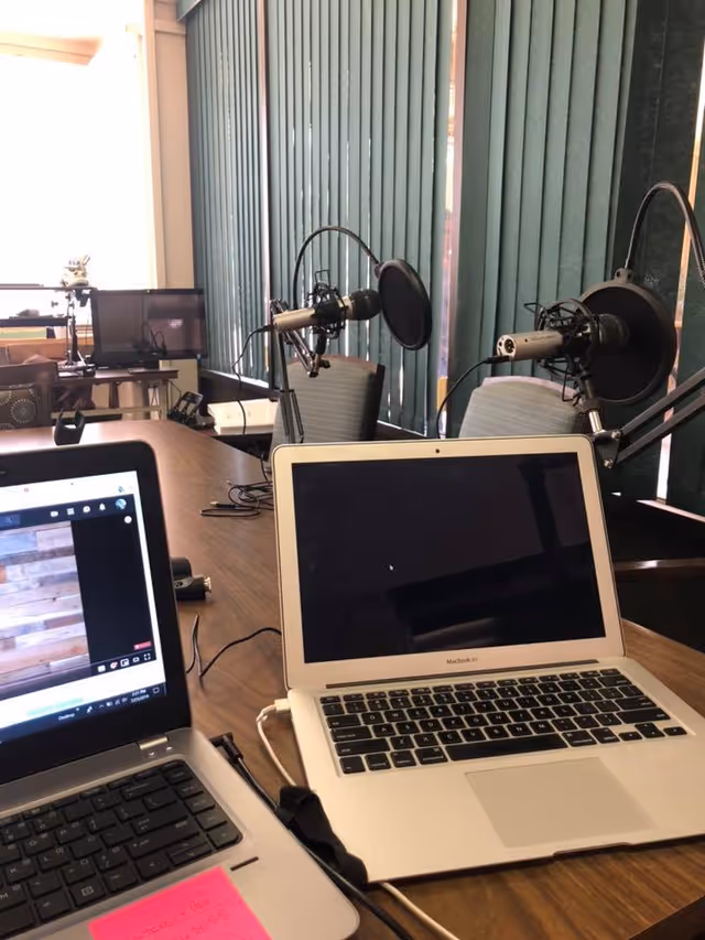Conference-style room table with two laptops and mounted podcast microphones against vertical blinds.