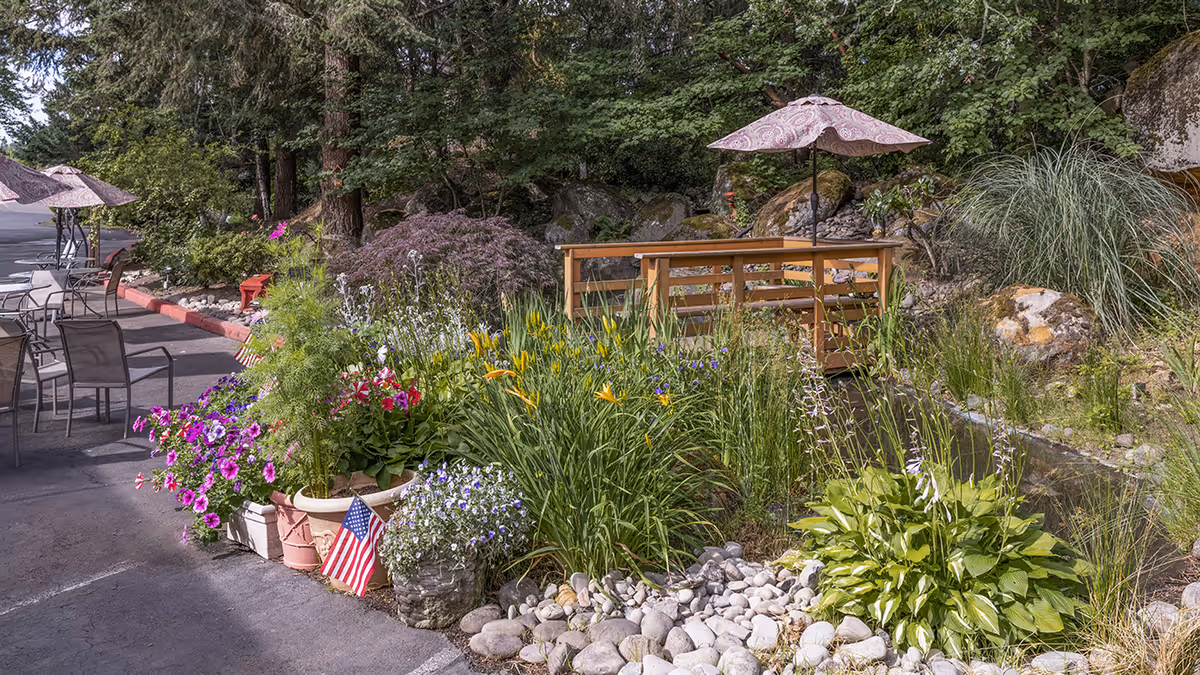 Outdoor garden patio area with potted flowers, a small wooden bridge over a pond and parasols near seating.