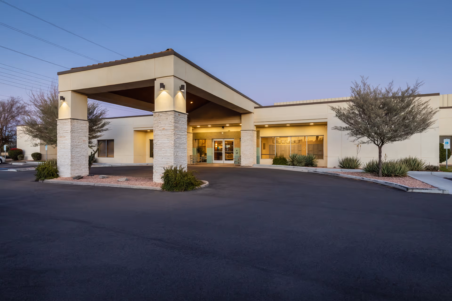 Exterior view of BridgeWater Assisted Living - Deer Valley building at dusk, showing the entrance with a covered drop-off area supported by stone pillars, surrounded by desert landscaping and a paved driveway.