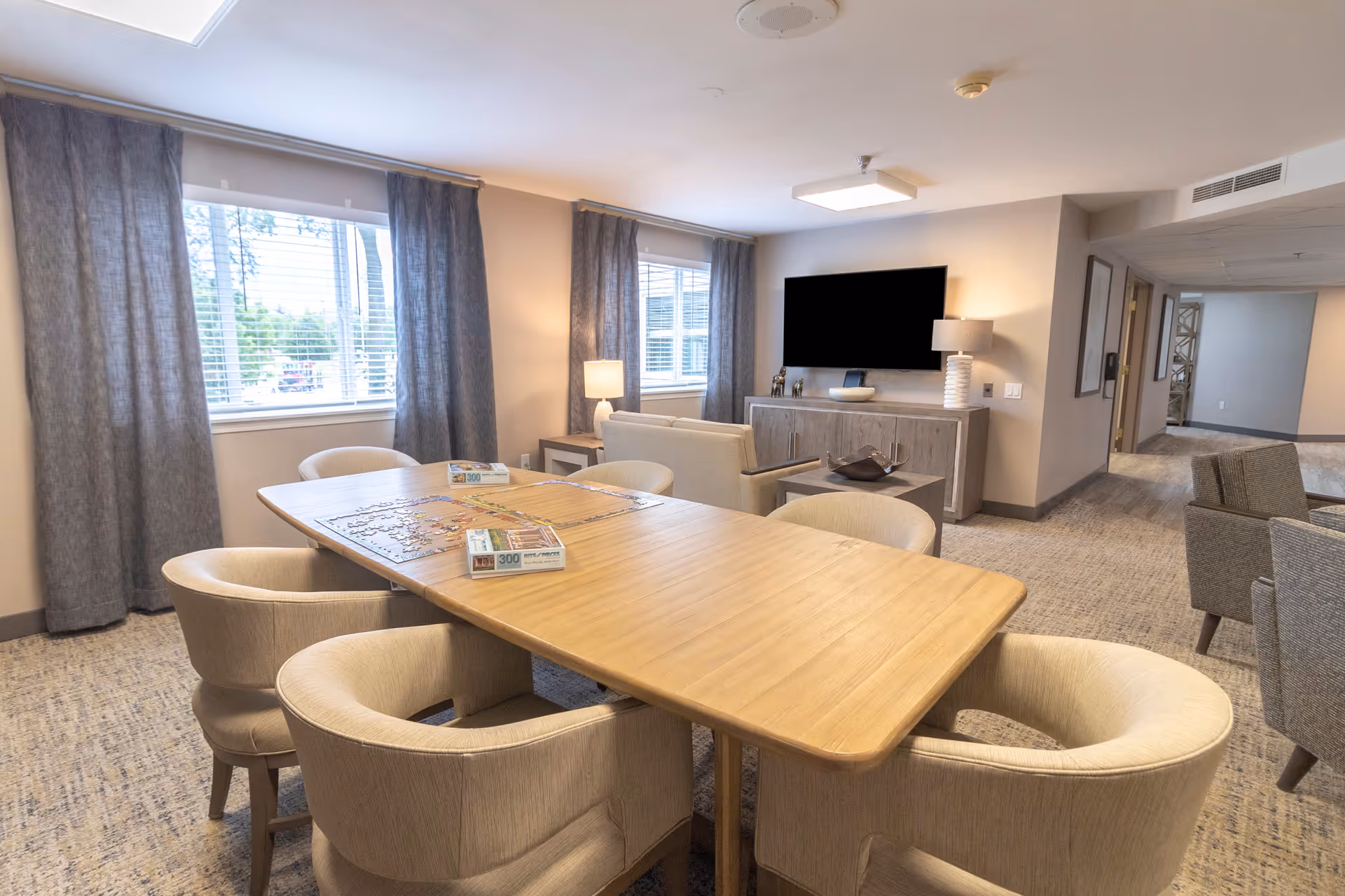 A well-lit common area with a large wooden table surrounded by six beige upholstered chairs. On the table, there are two puzzle boxes and a partially completed puzzle. Behind the table, there is a seating area with a sofa, a side table with a lamp, and a large flat-screen TV mounted on the wall above a wooden cabinet. The room has two windows with gray curtains, and the floor is carpeted in a neutral tone. A hallway is visible to the right.