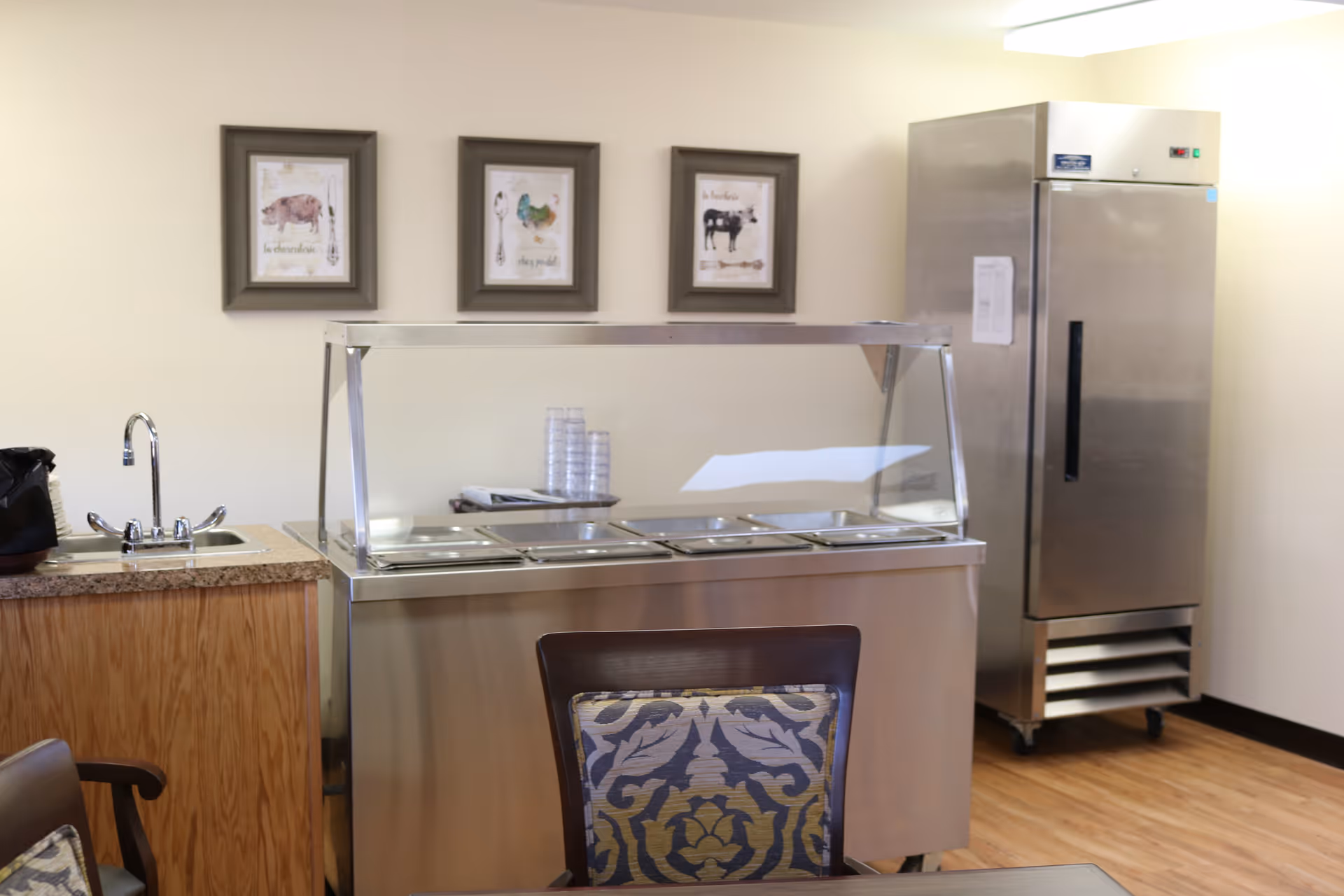 Small dining/cafeteria serving area with a stainless-steel food service counter, upright refrigerator, sink, and framed art on the wall.