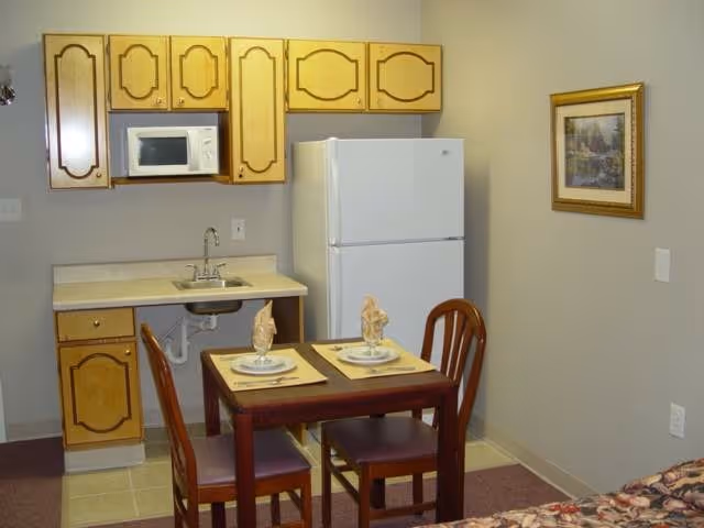 Small kitchenette with wooden cabinets, a sink, white refrigerator, microwave and a two-person dining table with place settings.