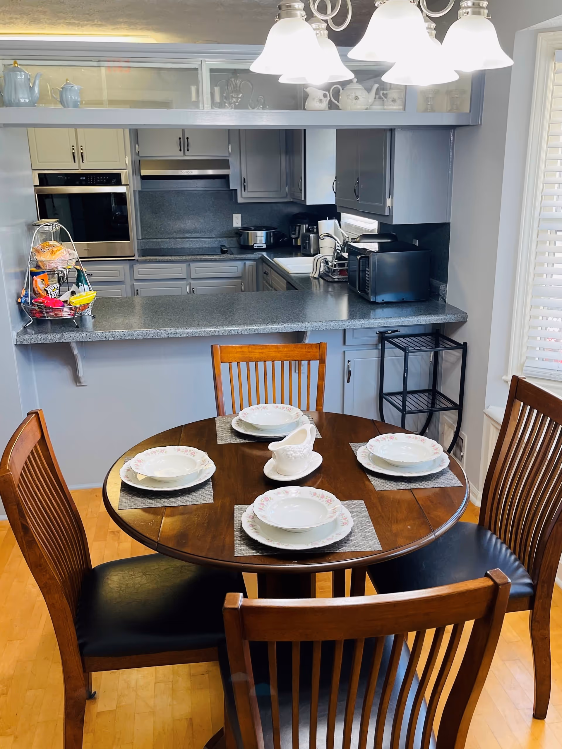 A dining area with a round wooden table set for four with floral-patterned plates and bowls, and a white gravy boat in the center. Four wooden chairs with black cushioned seats surround the table. Behind the table is a kitchen with gray cabinets, a countertop, a microwave, a slow cooker, and various kitchen appliances. A three-light chandelier hangs above the dining table.