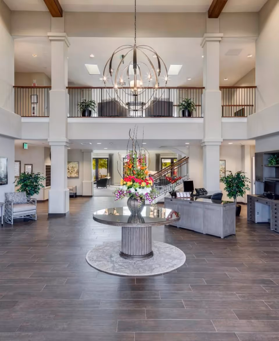 Spacious and well-lit lobby area with a round table in the center holding a large floral arrangement. The space features high ceilings with exposed wooden beams, a modern chandelier, and a second-floor balcony with seating and plants. There is a reception desk to the right and comfortable seating areas to the left and in the background.