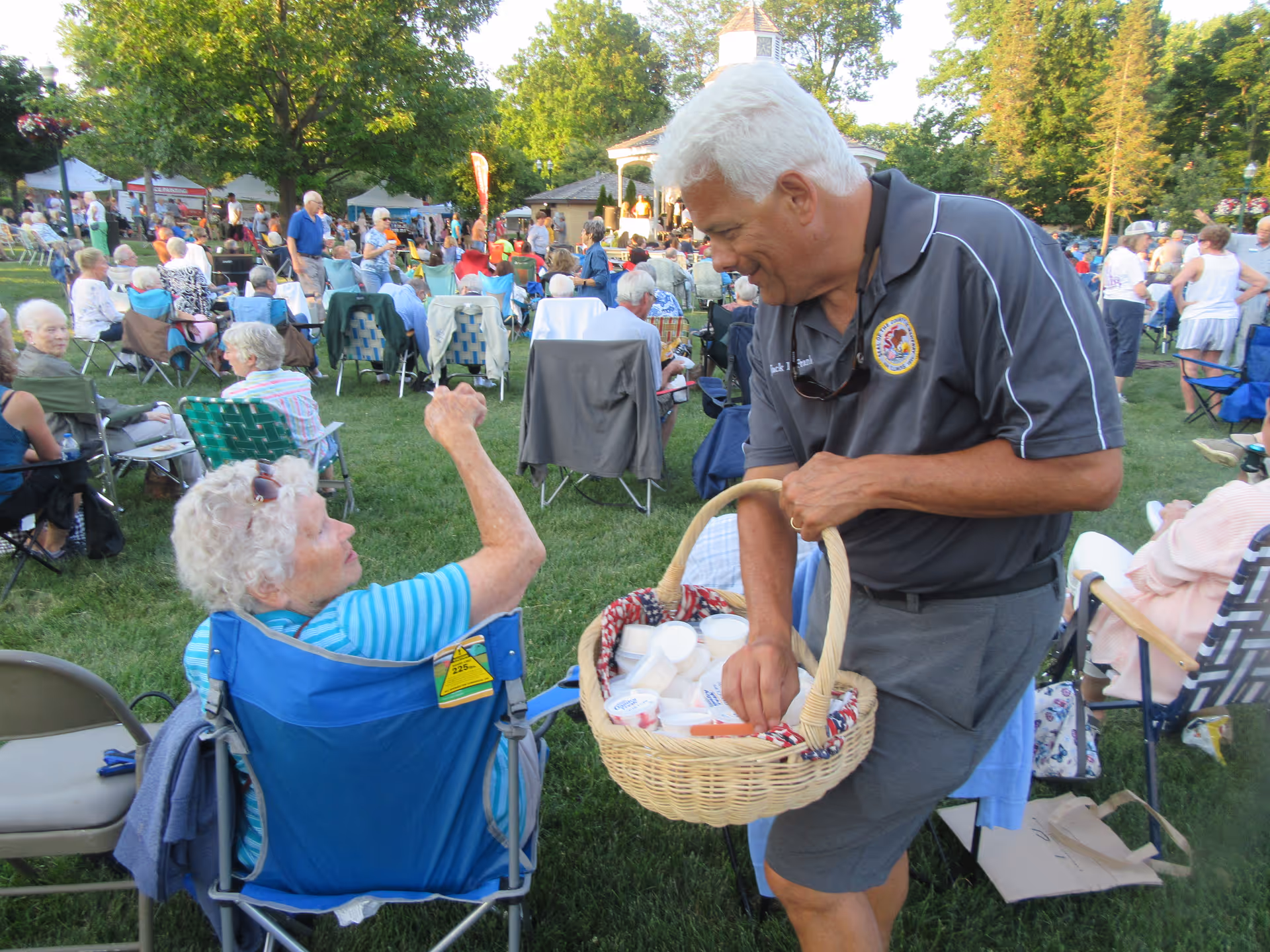 An outdoor gathering with many elderly people seated on lawn chairs on a grassy area. A man with white hair wearing a dark polo shirt and gray shorts is holding a wicker basket filled with cups and is interacting with an elderly woman seated in a blue chair. Trees and a gazebo are visible in the background.