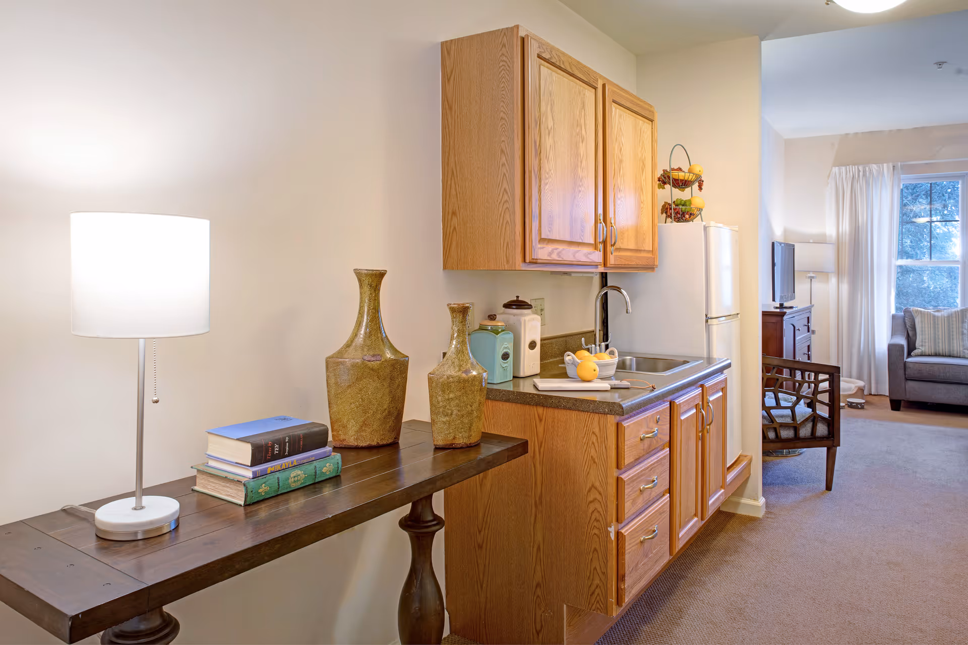 Interior view of a senior living facility room at Sunrise at Siegen featuring a small kitchenette with wooden cabinets, a sink, and a refrigerator. To the left, there is a wooden table with a lamp, two decorative vases, and a stack of books. In the background, a living area is visible with a chair, a TV on a stand, a floor lamp, and a window with white curtains.