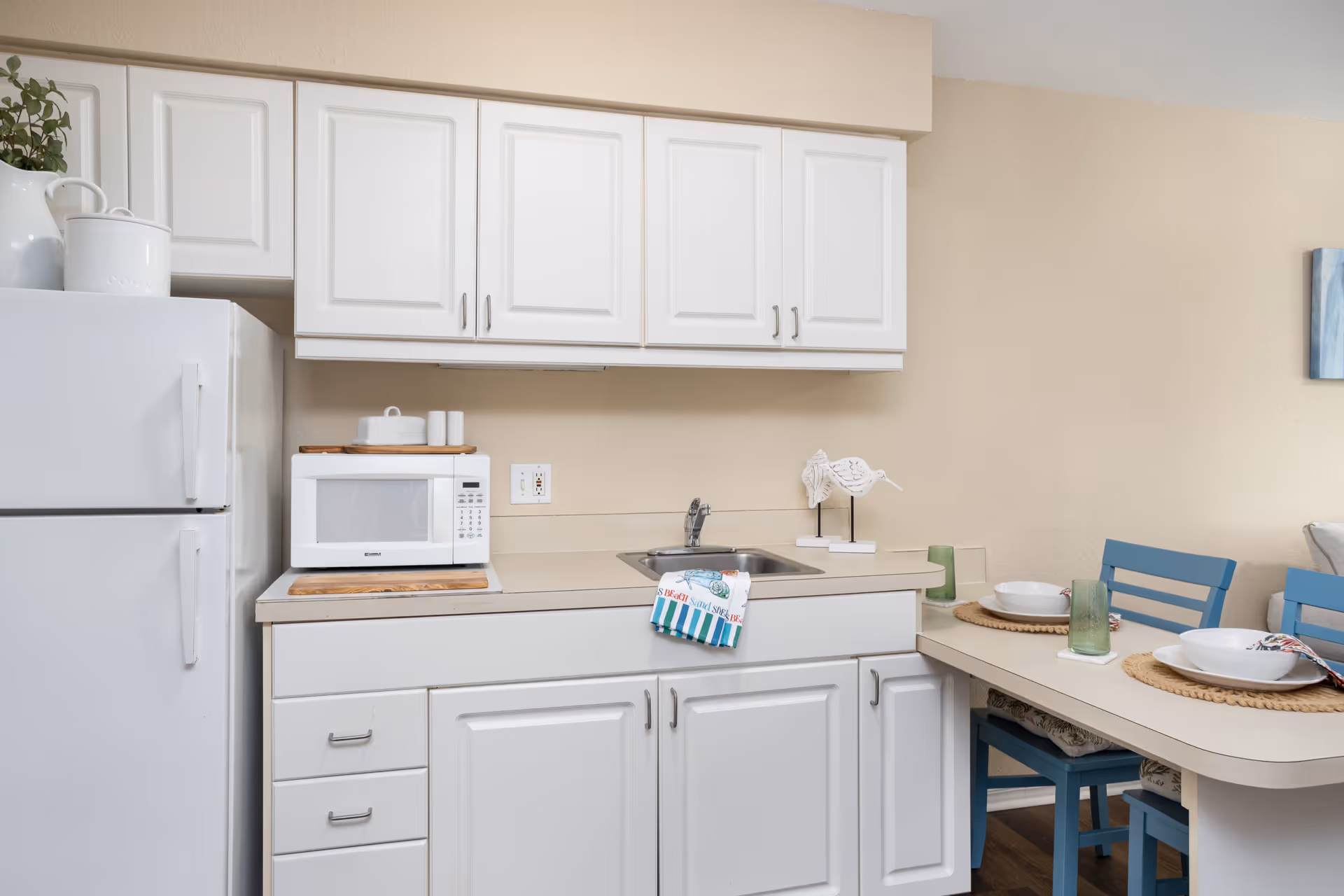A small kitchen area with white cabinets, a white refrigerator, a microwave on the countertop, a small sink, and a dining counter with two place settings including bowls, glasses, and napkins. There are two blue chairs at the dining counter.