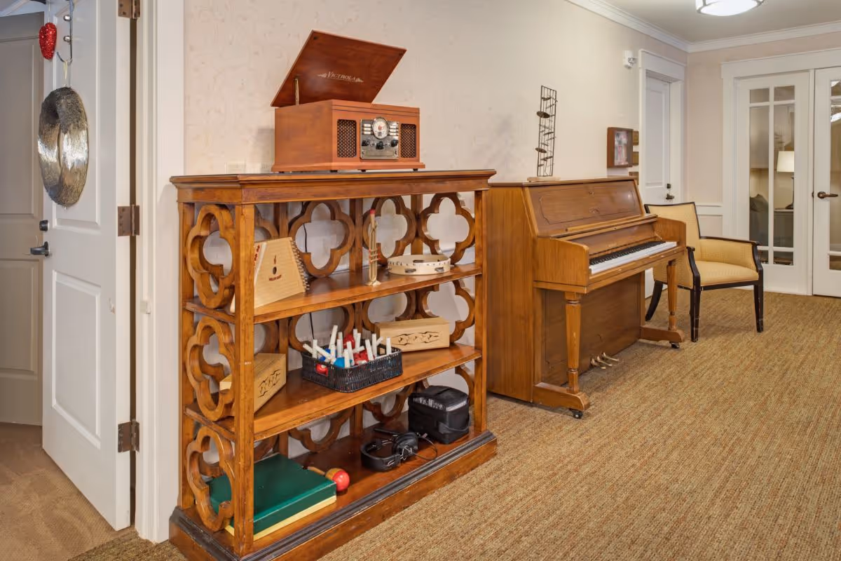 Upright wooden piano beside a decorative wooden shelf holding small musical instruments and a chair in a carpeted common area.