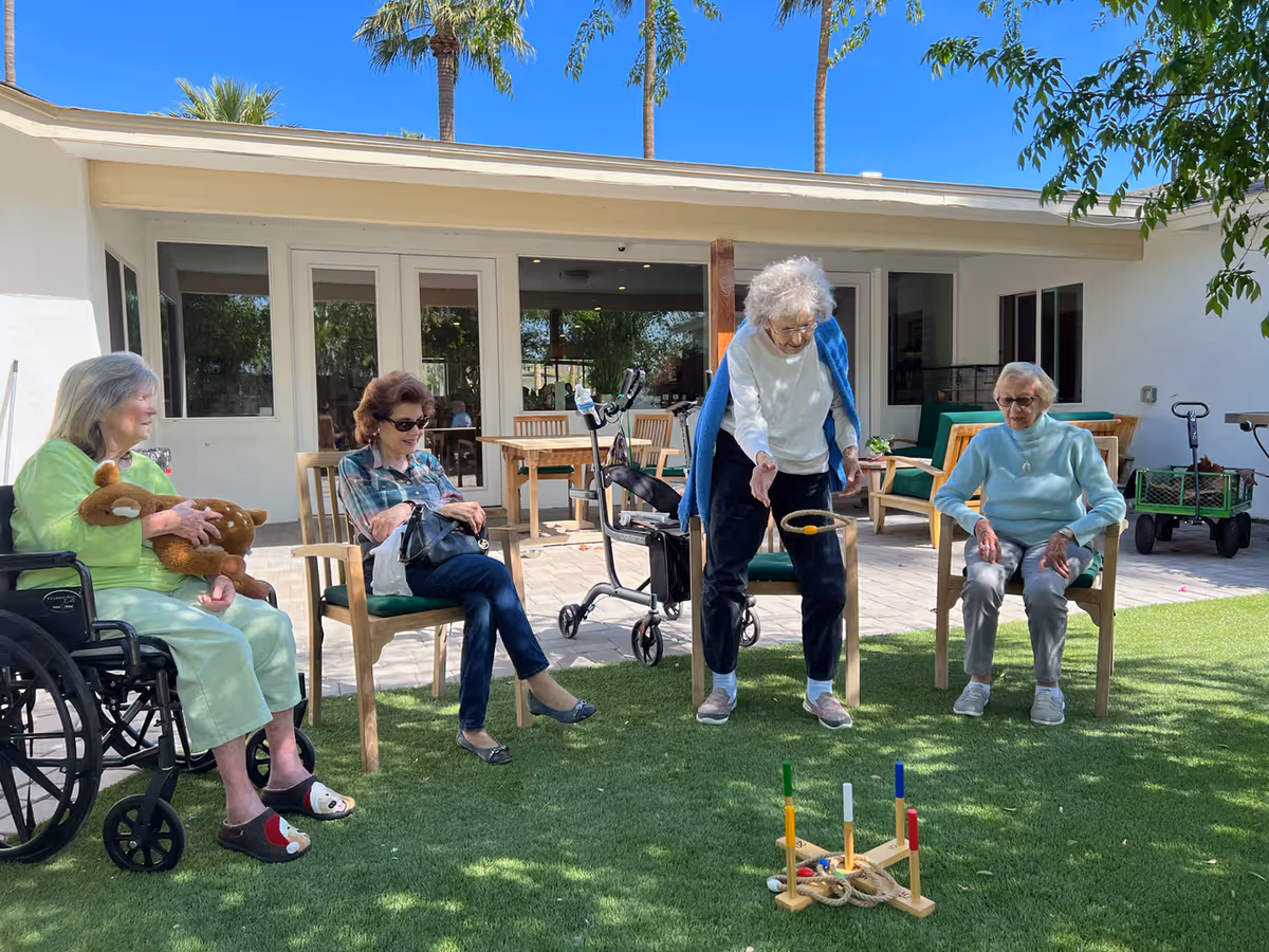 Four elderly women seated outside a senior living facility play a ring toss game on a grassy courtyard in front of a building.
