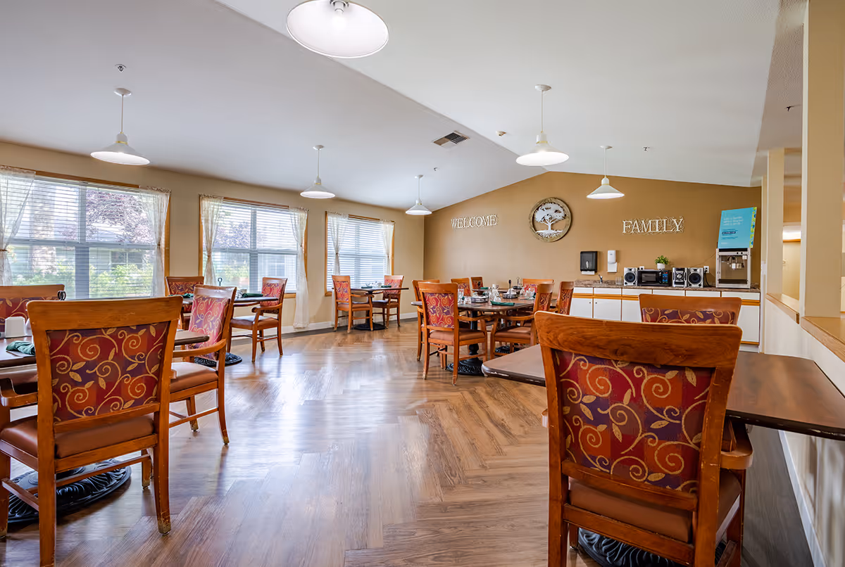 Communal dining room with wooden tables and patterned chairs, large windows, and a serving counter under wall décor reading 'WELCOME' and 'FAMILY'.