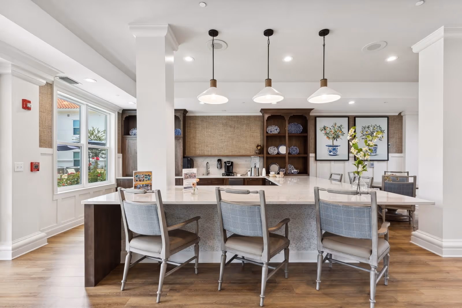 A bright and modern kitchen area with a large marble countertop island surrounded by four cushioned chairs. Three pendant lights hang above the island. Behind the island, there are wooden cabinets with decorative blue and white plates and two framed botanical prints on the wall. A window on the left side lets in natural light, and a vase with flowers is placed on the island.