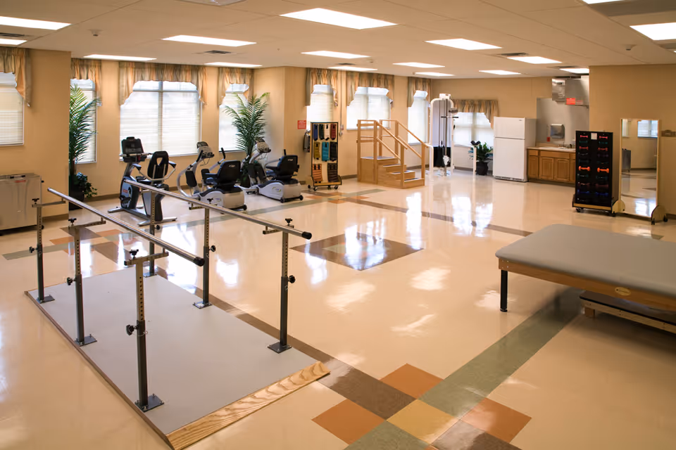 A spacious physical therapy or rehabilitation room with exercise equipment including stationary bikes, parallel bars for walking practice, a wooden staircase for step exercises, a treatment table, and racks holding various weights. The room has large windows with curtains, beige walls, and a tiled floor with a colorful pattern.