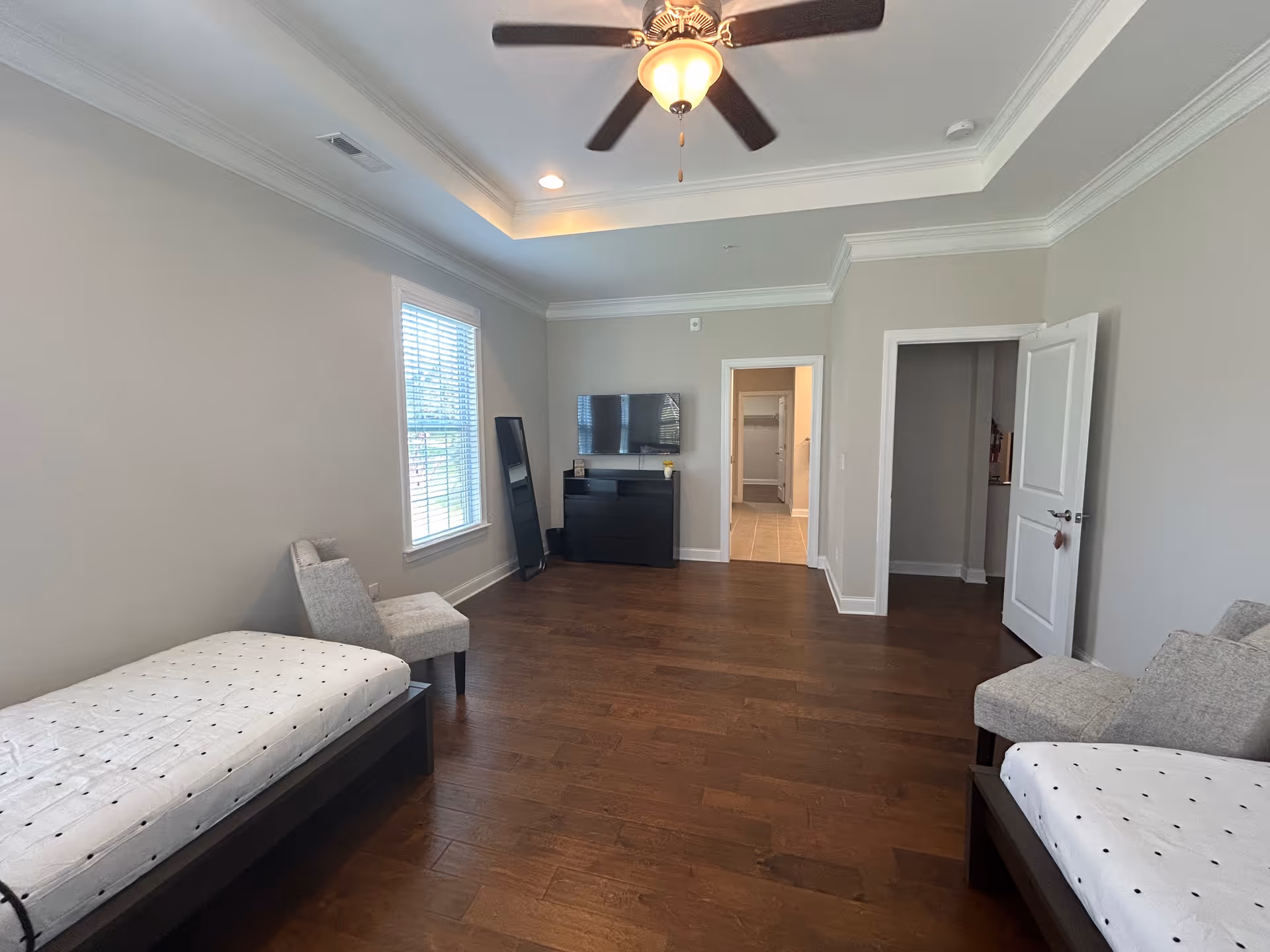 A spacious bedroom with two twin beds, each with white bedding featuring small black dots. There are two gray upholstered chairs, one near a window with white blinds and the other near the foot of one bed. A black dresser with a flat-screen TV mounted above it is against the far wall. The room has hardwood floors, light gray walls, a ceiling fan with a light fixture, and two open doorways leading to other rooms.