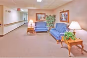 Carpeted interior hallway with two blue upholstered sofas, wooden side tables with lamps, framed artwork, and a potted plant.