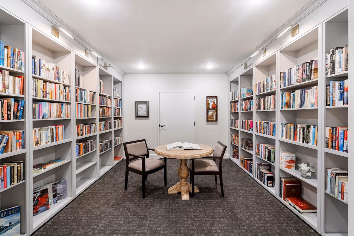 A small library room with white built-in bookshelves filled with books on both sides. In the center, there is a round wooden table with an open book on it and two cushioned chairs facing each other. The room has a carpeted floor, white walls, and a closed white door at the back. A clock and a framed picture hang on the wall beside the door.
