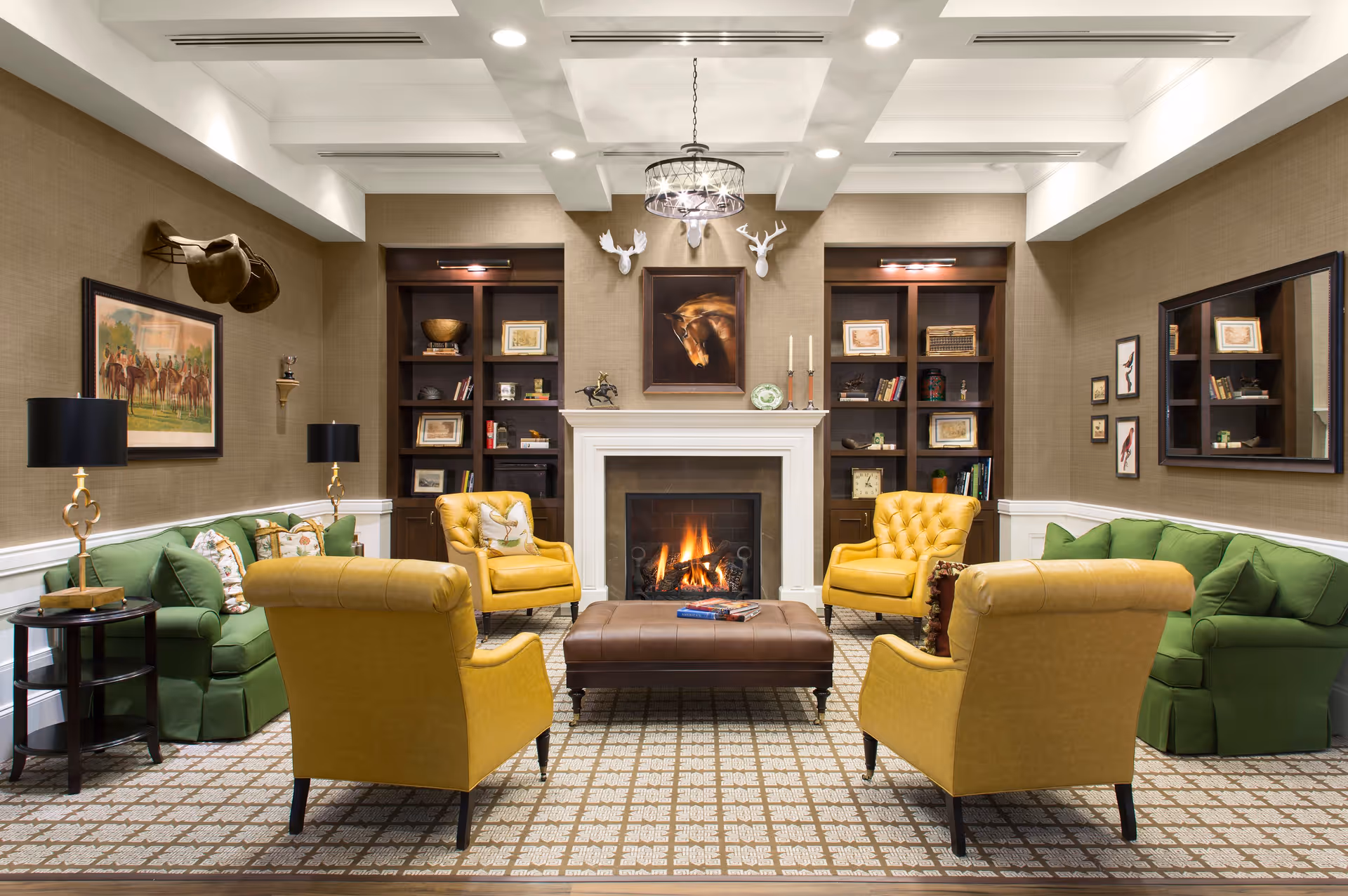 A cozy living room with a lit fireplace in the center, flanked by two dark wooden bookshelves filled with books and decorative items. The room features two green sofas on either side and four yellow armchairs arranged around a large brown ottoman in the middle. The walls are decorated with framed artwork, including a horse painting above the fireplace and mounted deer heads. The ceiling has recessed lighting and a decorative chandelier.