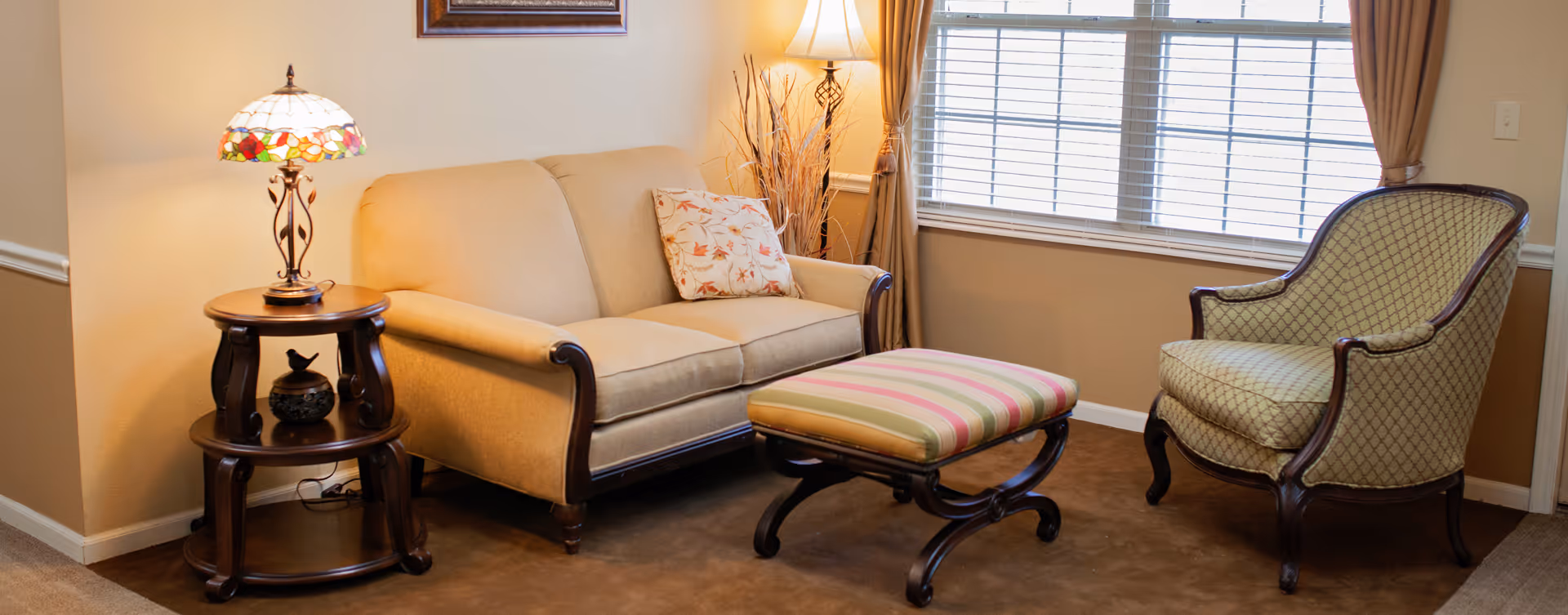 A cozy living room area with a beige sofa, a patterned armchair, and a striped ottoman. There is a wooden side table with a decorative lamp and a small figurine. A window with blinds and beige curtains allows natural light into the room.