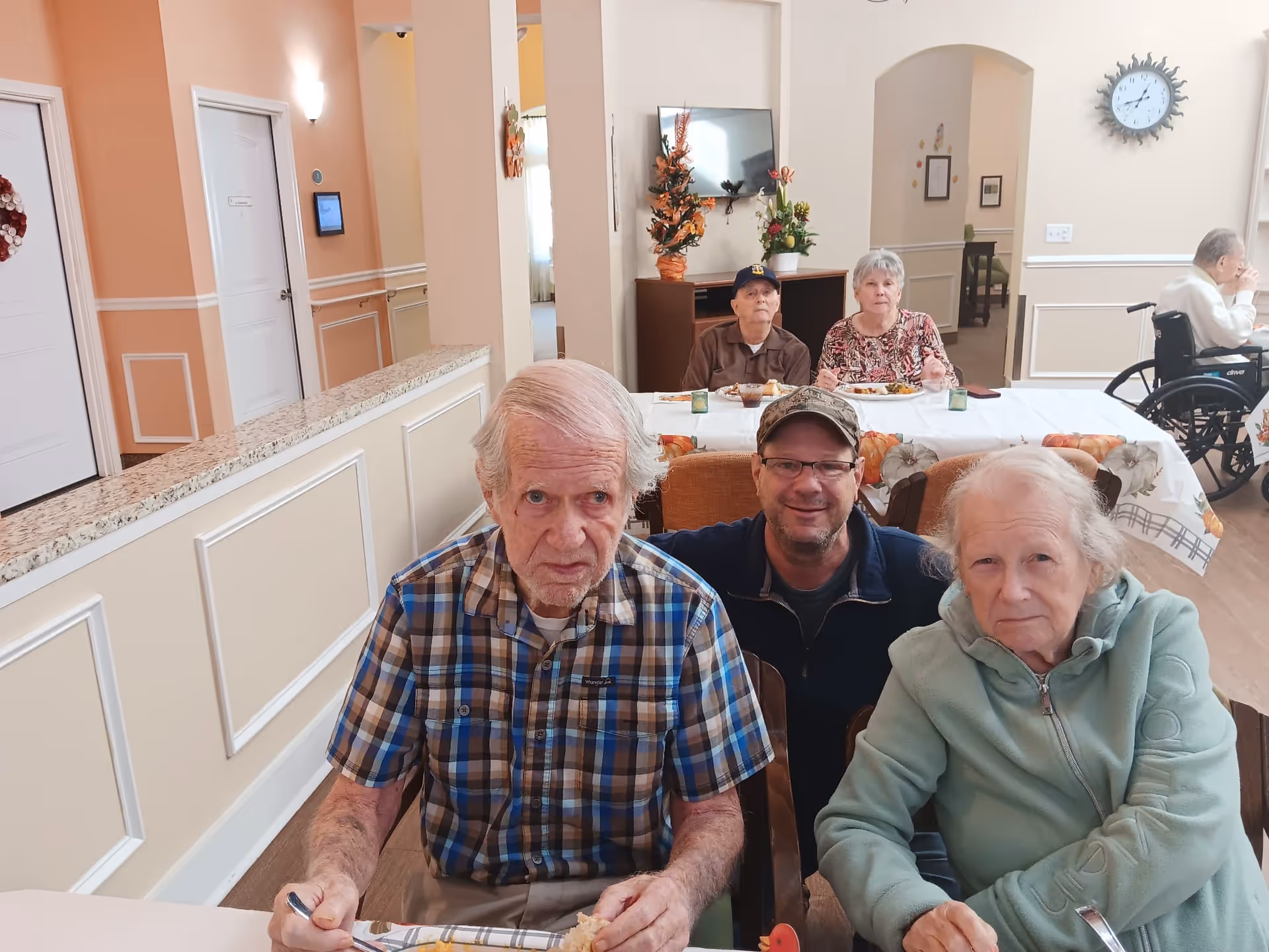 A group of elderly people and a middle-aged man sitting together at a dining table in a well-lit room with peach-colored walls. The table is decorated with a fall-themed tablecloth and small pumpkins. In the background, more elderly people are seated at another table, and one person is in a wheelchair. The room has a clock on the wall and a TV mounted above a cabinet with floral decorations.