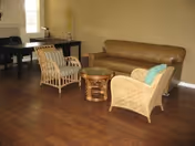 A cozy living room area with wooden flooring, featuring a brown leather sofa, two wicker armchairs with cushions, a small round wooden coffee table, and a black desk with a chair near a window.