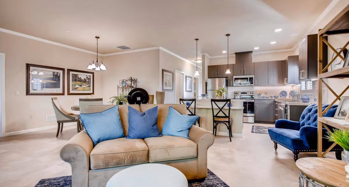 A bright and spacious open-concept living area featuring a beige sofa with blue cushions, a round white coffee table, a blue armchair, and a wooden shelving unit with decorative items. In the background, there is a dining area with a table and chairs, and a modern kitchen with dark cabinetry, stainless steel appliances, and pendant lighting.